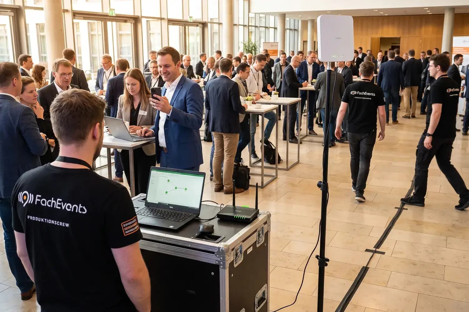 Foyer einer Konferenz in der Pause: Gäste mit Smartphones und Laptops, ein WLAN-Access-Point auf einem Stativ, vorn ein Techniker am Netzwerkplatz
